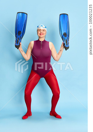 Happy swimmer. Full-length image of woman in swimming suit and red tights posing with flippers against blue studio background 100021235