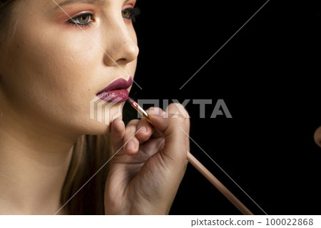 Makeup artist applies red lipstick on a beautiful woman face. Hand of make-up master, painting lips of young beauty model girl. Make up in process. Makeup artist applies red lipstick on a beautiful woman face. Hand of make-up master, painting lips of young beauty model girl. Make up in process. 100022868