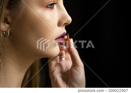 Makeup artist applies red lipstick on a beautiful woman face. Hand of make-up master, painting lips of young beauty model girl. Make up in process. 100022907