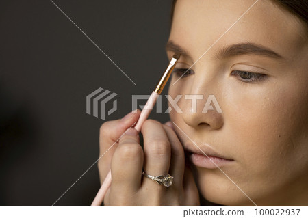 Makeup artist doing a eyebrows makeup on a beautiful woman face. Hand of make-up master is painting eyebrow of young beauty model girl. Make up in process. Makeup artist doing a eyebrows makeup on a beautiful woman face. Hand of make-up master is painting eyebrow of young beauty model girl. Make up in process. 100022937