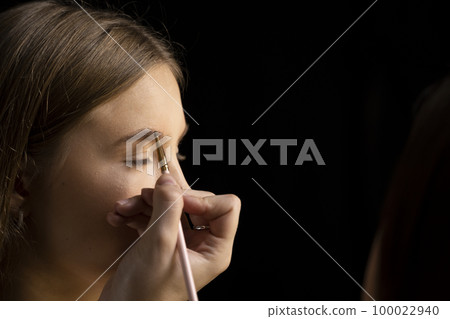 Makeup artist doing a eyebrows makeup on a beautiful woman face. Hand of make-up master is painting eyebrow of young beauty model girl. Make up in process. 100022940
