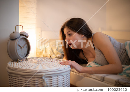 A girl with long hair in shock checks the time on her alarm clock. A girl with long hair in shock checks the time on her alarm clock. 100023610