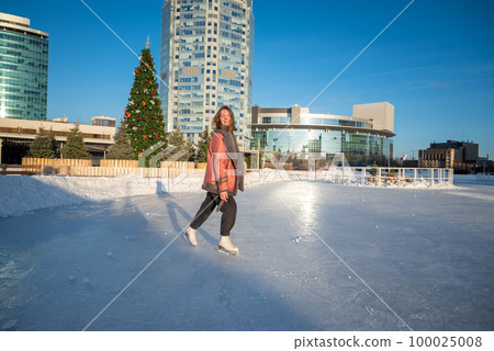 a young beautiful girl on skates in a sheepskin coat on a skating rink against the backdrop of the metropolis and the rays of the sun 100025008
