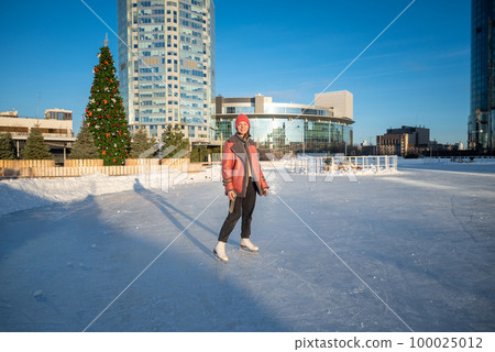 a young beautiful girl on skates in a sheepskin coat on a skating rink against the backdrop of the metropolis and the rays of the sun 100025012