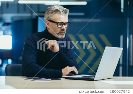 Thinking businessman inside the office with a laptop, mature adult boss thinking about the decision for working sitting with a computer. Thinking businessman inside the office with a laptop, mature adult boss thinking about the decision for working sitting with a computer. 100027942