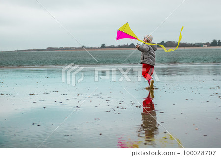 Happy child is running with bright colouful kite on the sea beach. Kid girl flying the kite outdoors the coast shore. Reflection in water 100028707