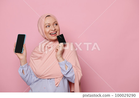 Middle-aged Arab Muslim pensive woman holding a smartphone and blank plastic credit card, thoughtfully looking aside at copy space, isolated pink background. Shopping online. Ordering food. Booking 100029290