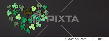 Composition made of knitted green shamrocks in the shape of a heart on a black background. Holiday sign and knitted clover leaf. Copy space, flat lay, banner. St Patrick's Day concept 100030410
