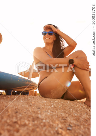 Charming woman with long hair and sporty tanned body smiling sitting in the setting sun with a surfboard on the beach on a sunny summer day, ready to surf. in a bathing suit 100032708