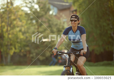 Smiling middle aged woman in cap with glasses cycling in summer park, cycling cardio training, city walks with bike, weight control Smiling middle aged woman in cap with glasses cycling in summer park, cycling cardio training, city walks with bike, weight control 100033025