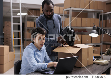 Storehouse employee putting customer helmet order in carton box preparing delivery for client while discussing transportation details with manager. Diverse team working in warehouse 100038920