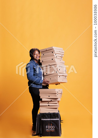 Smiling deliverywoman holding stack full of pizza boxes preparing to deliver to clients during lunch time. Restaurant emloyee with takeout thermal backpack delivering takeaway food order Smiling deliverywoman holding stack full of pizza boxes preparing to deliver to clients during lunch time. Restaurant emloyee with takeout thermal backpack delivering takeaway food order 100038986