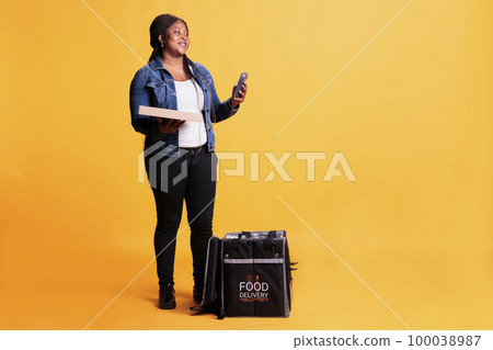 Deliverywoman holding carton box full with pizza while checking client adreess on phone using takeaway food app before delivery food order. Employee standing in studio with yellow background Deliverywoman holding carton box full with pizza while checking client adreess on phone using takeaway food app before delivery food order. Employee standing in studio with yellow background 100038987