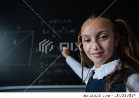 Portrait of a caucasian girl in the classroom. The schoolgirl writes the formula with chalk on the blackboard and looks into the camera. 100039814