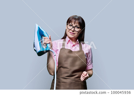 Woman in apron posing with iron, on gray background 100045936