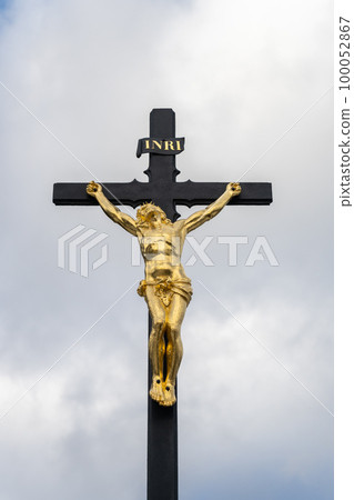 Golden statue of crucified Jesus with a Latin inscription INRI, standing on an iron cross, against a cloudy background. 100052867