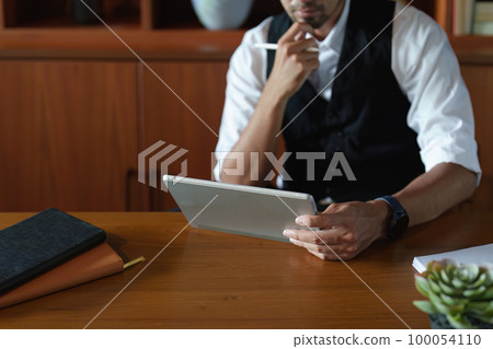 A portrait of a good looking and discreet Asian man sitting at his desk with a thoughtful look on his tablet computer. A portrait of a good looking and discreet Asian man sitting at his desk with a thoughtful look on his tablet computer. 100054110
