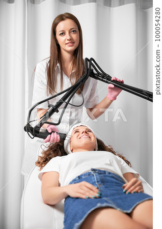 Visiting beautician to treat post-traumatic acne scars. Female dermatologist performing laser resurfacing on face skin of smiling young woman. 100054280