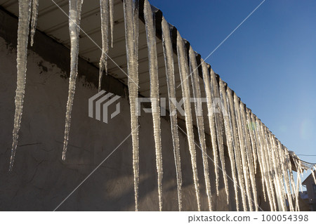 Icicles hang on the roof of a house against a bright blue sky. Spring landscape with icicles hanging from the roof of the house. Set of snow icicles, snow hat Icicles hang on the roof of a house against a bright blue sky. Spring landscape with icicles hanging from the roof of the house. Set of snow icicles, snow hat 100054398