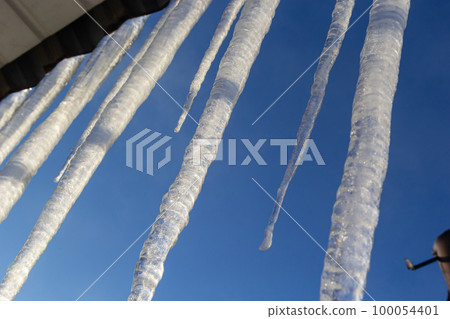 Icicles hang on the roof of a house against a bright blue sky. Spring landscape with icicles hanging from the roof of the house. Set of snow icicles, snow hat Icicles hang on the roof of a house against a bright blue sky. Spring landscape with icicles hanging from the roof of the house. Set of snow icicles, snow hat 100054401