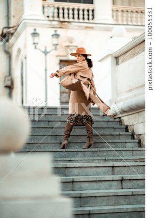 Outdoor fashion portrait of young elegant fashionable brunette woman, model in stylish hat, choker and light raincoat posing at sunset in European city. Outdoor fashion portrait of young elegant fashionable brunette woman, model in stylish hat, choker and light raincoat posing at sunset in European city. 100055331