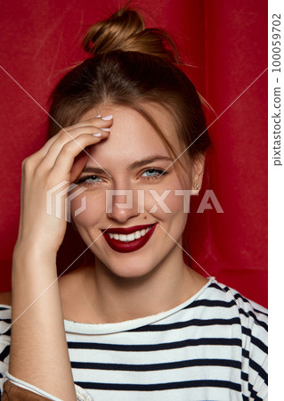 Positive femininity. Portrait of young beautiful girl with red lips and hair bun, in striped shirt posing against red studio background. Concept of beauty and fashion Positive femininity. Portrait of young beautiful girl with red lips and hair bun, in striped shirt posing against red studio background. Concept of beauty and fashion 100059702