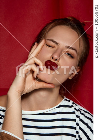 Portrait of young beautiful girl with red lips and hair bun, in striped shirt posing against red studio background. Parisian style. Concept of beauty and fashion Portrait of young beautiful girl with red lips and hair bun, in striped shirt posing against red studio background. Parisian style. Concept of beauty and fashion 100059703