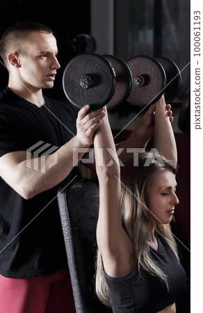 Young woman with personal trainer exercising on the simulator in the gym 100061193