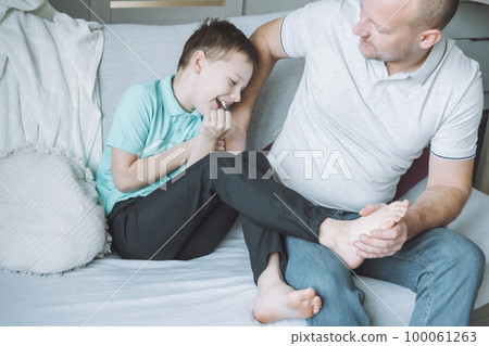 Father plays with his son 7-10 sitting on couch. Dad tickles kids feet. Family, having fun 100061263