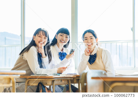 一名高中生在教室裡看著他的智能手機與他的同學交談 一名高中生在教室裡看著他的智能手機與他的同學交談 100062773