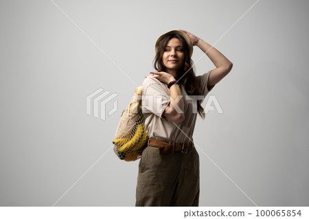 Beautiful brunette woman in beige t-shirt and black hat holding cotton reusable mesh shopping bags with groceries, pepper, broccoli. Zero waste, plastic free concept. 100065854