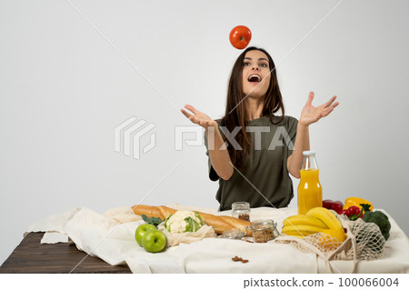 Happy woman in green t-shirt unpacking shopping mesh eco bag with healthy vegetables, fruits, bread, snacks on the kitchen at home. Healthy eating vegetarian concept. 100066004