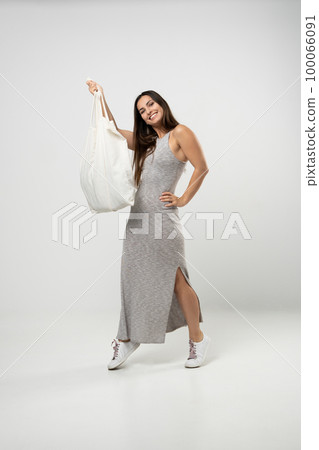 Brunette woman in grey dress holding cotton shopper bag with vegetables, products in white room. Eco friendly shopping bags. Zero waste, plastic free concept. Brunette woman in grey dress holding cotton shopper bag with vegetables, products in white room. Eco friendly shopping bags. Zero waste, plastic free concept. 100066091