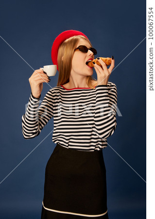 Portrait of beautiful young girl in red beret posing, eating croissant with coffee over blue studio background. Concept of fashion, beauty and lifestyle 100067554