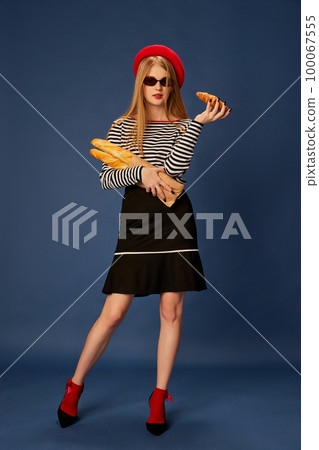 Portrait of beautiful young girl in red beret posing with baguette and croissant over blue studio background. Concept of fashion, beauty and lifestyle Portrait of beautiful young girl in red beret posing with baguette and croissant over blue studio background. Concept of fashion, beauty and lifestyle 100067555
