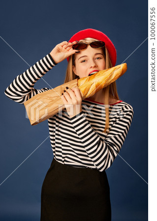 Parisian style. Portrait of beautiful young girl in red beret posing, biting baguette over blue studio background. Concept of fashion, beauty and lifestyle Parisian style. Portrait of beautiful young girl in red beret posing, biting baguette over blue studio background. Concept of fashion, beauty and lifestyle 100067556