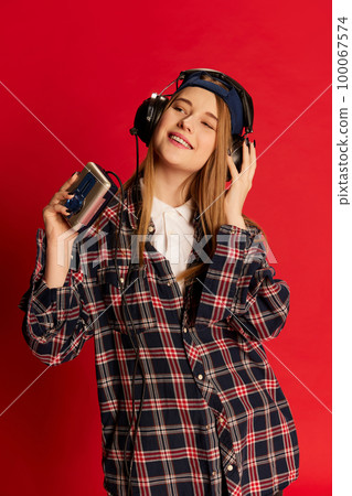 Young beautiful positive girl in checkered shirt and cap listening to music in headphones with player over red studio background. Concept of lifestyle and youth 100067574
