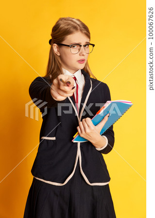 Time to study. Portrait of beautiful young girl, student in uniform with notes pointing with pencil over yellow studio background. Concept of emotions, youth, education and lifestyle 100067626