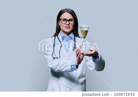 Young woman doctor holding hourglass, on light gray background Young woman doctor holding hourglass, on light gray background 100072650