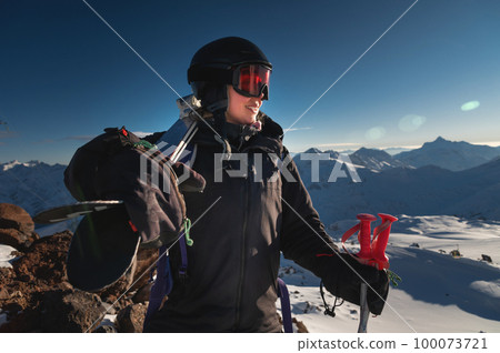 Image of a woman with skis on her shoulder against a snowy hill or mountain range. Portrait of a cheerful skier carrying skis on her shoulder 100073721