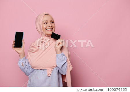 Delightful smiling Muslim woman in pink hijab, looking aside, holding a smartphone and plastic bank credit card, ordering food delivery service, shopping online or booking tour via mobile application Delightful smiling Muslim woman in pink hijab, looking aside, holding a smartphone and plastic bank credit card, ordering food delivery service, shopping online or booking tour via mobile application 100074365