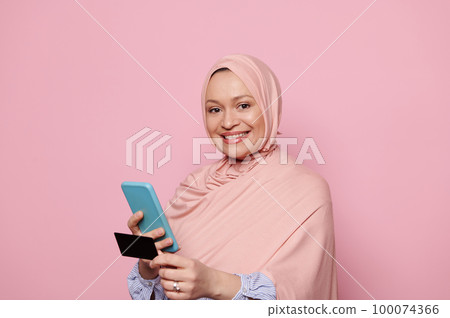 Muslim woman in pink hijab, holding smartphone and credit card, online shopping, provide internet payment. Happy client enjoys easy fast secure transferring money in bank app. Safe mobile banking. Muslim woman in pink hijab, holding smartphone and credit card, online shopping, provide internet payment. Happy client enjoys easy fast secure transferring money in bank app. Safe mobile banking. 100074366