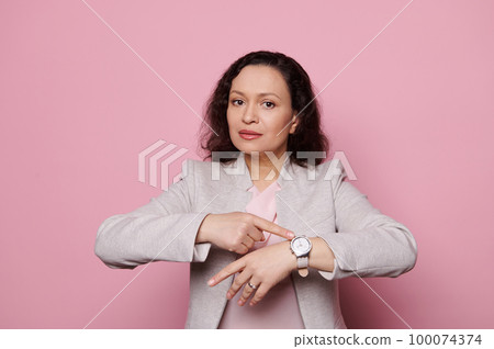 Attractive multi-ethnic businesswoman in elegant light gray formal jacket, showing you the time on her wristwatch, confidently looking at camera, isolated pink background. People. Punctuality concept 100074374