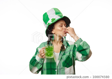 Happy woman in party hat with clover leaves, kissing fingers, showing a delicious sign, enjoying the taste of a green beer, celebrating St. Patrick's Day in a pub, isolated white background. March 17 100074380