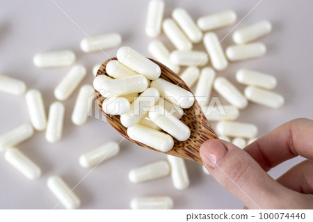 White vitamin C capsules in a wooden spoon on a beige background with scattered medications. White vitamin C capsules in a wooden spoon on a beige background with scattered medications. 100074440