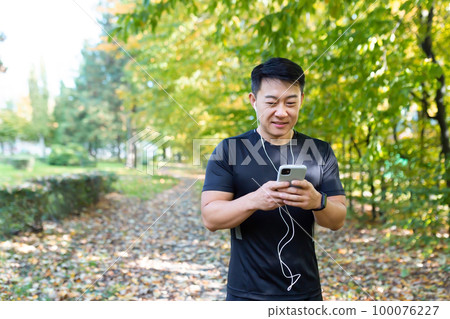 A young Asian male athlete is doing a morning jog in the park. He holds the phone in his hands, listens to music in headphones. 100076227
