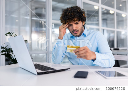 Problems with the account, bankruptcy, etc. A young hispanic man is sitting at the desk in the office, looking worriedly at the credit card, holding his head. 100076228