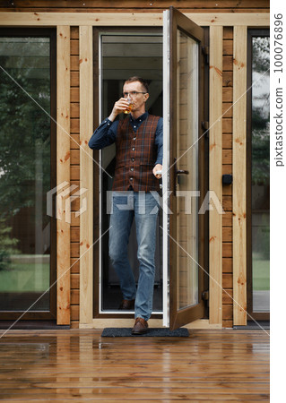 Middle age man comes out on the terrace of a small wooden house with a glass in hand Middle age man comes out on the terrace of a small wooden house with a glass in hand 100076896