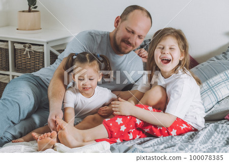 Father plays with two children on bed. Dad tickles kids feet. Family of daddy, and two little girls 100078385