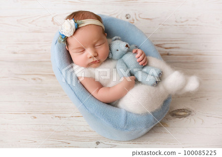 Top view of a newborn baby girl sleeping in a white jumpsuit with a blue teddy bear, in a blue felt chair. On a light wooden background. Beautiful portrait of a little girl 7 days, one week. Top view of a newborn baby girl sleeping in a white jumpsuit with a blue teddy bear, in a blue felt chair. On a light wooden background. Beautiful portrait of a little girl 7 days, one week. 100085225